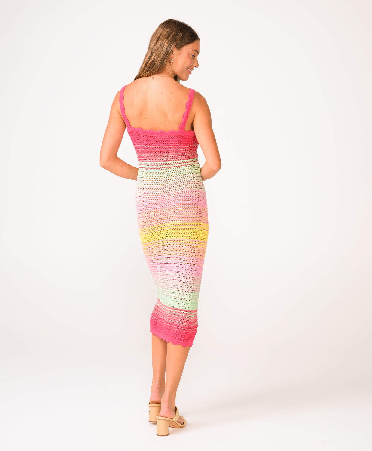 A woman wearing a pink, green, and yellow crochet knit mid length dress facing towards a white background.