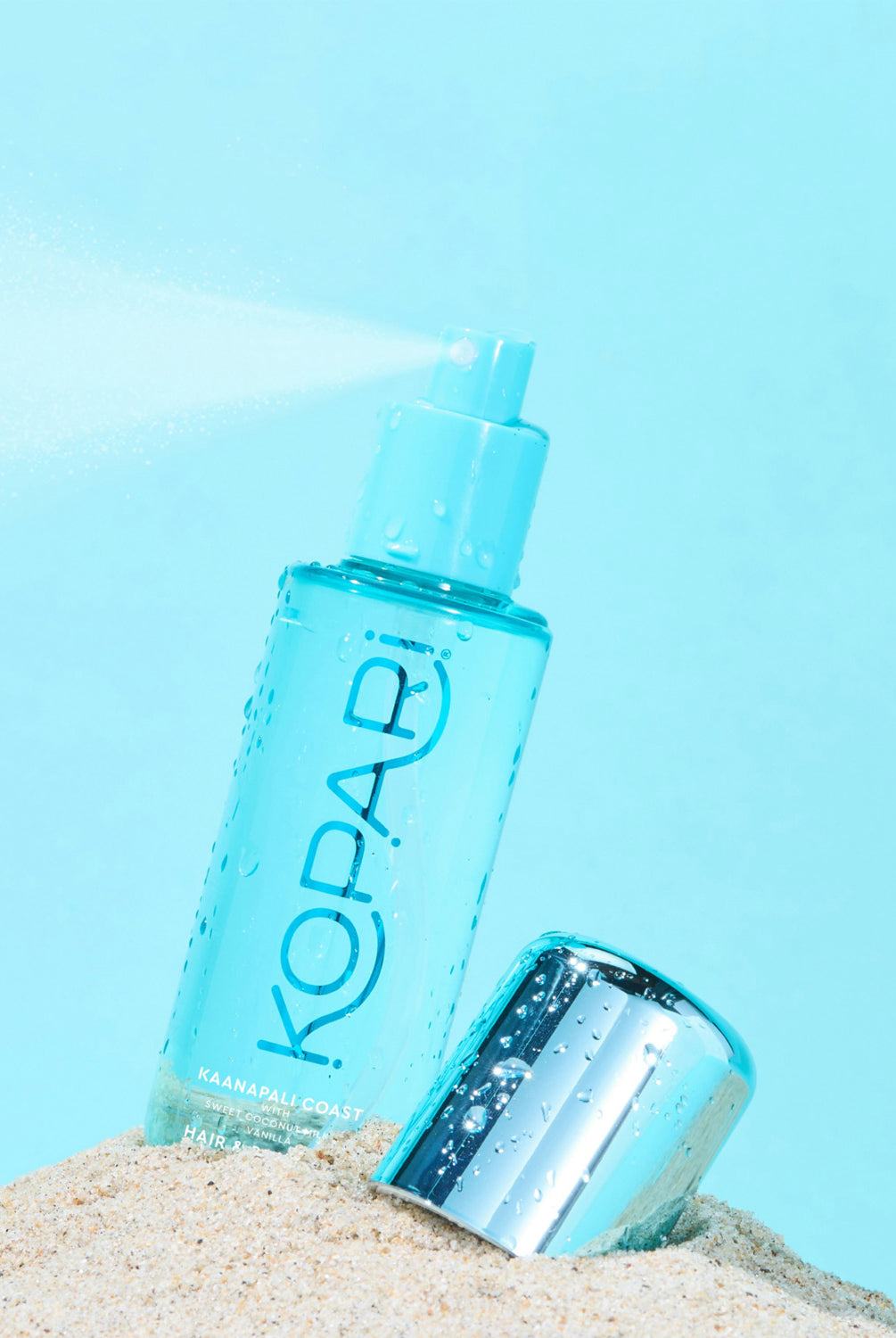 The Hair & Body Mist bottle resting in the sand with a bright blue sky backdrop.