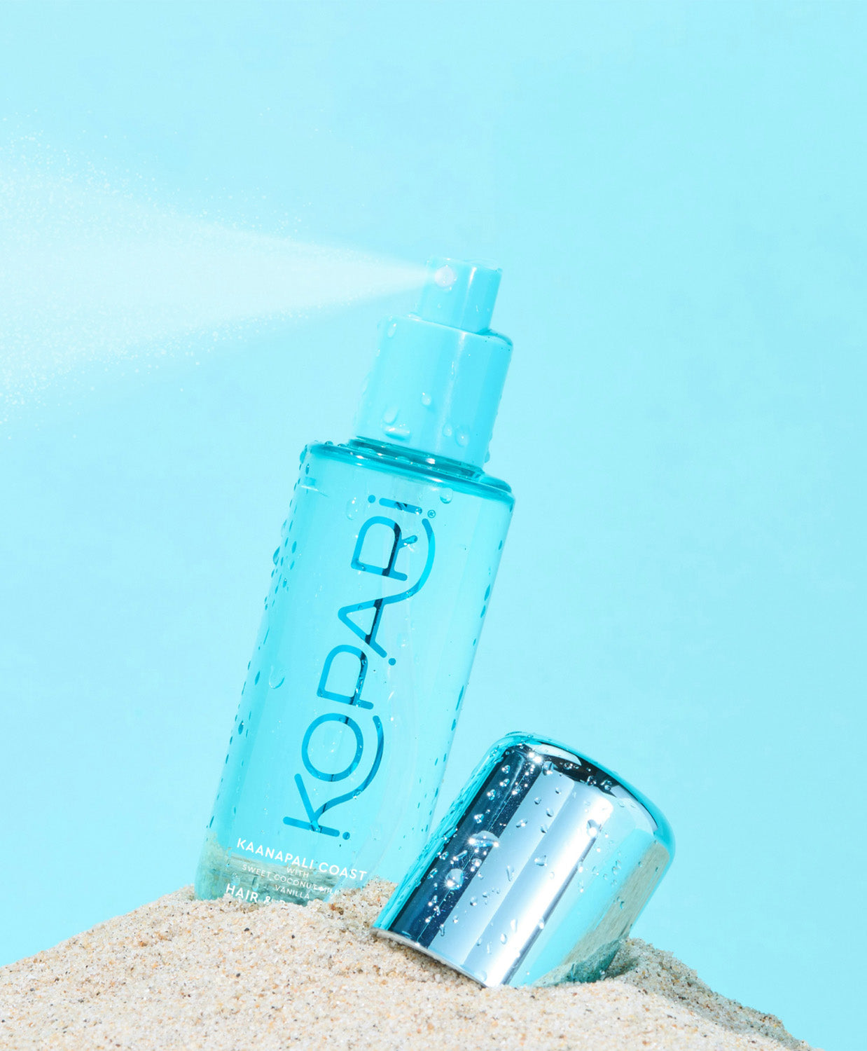The Hair & Body Mist bottle resting in the sand with a bright blue sky backdrop.