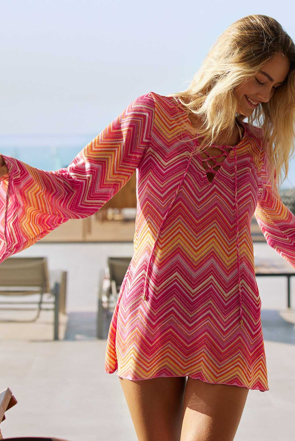 A woman wearing a pink and orange stripe long sleeve dress standing outside near the ocean.