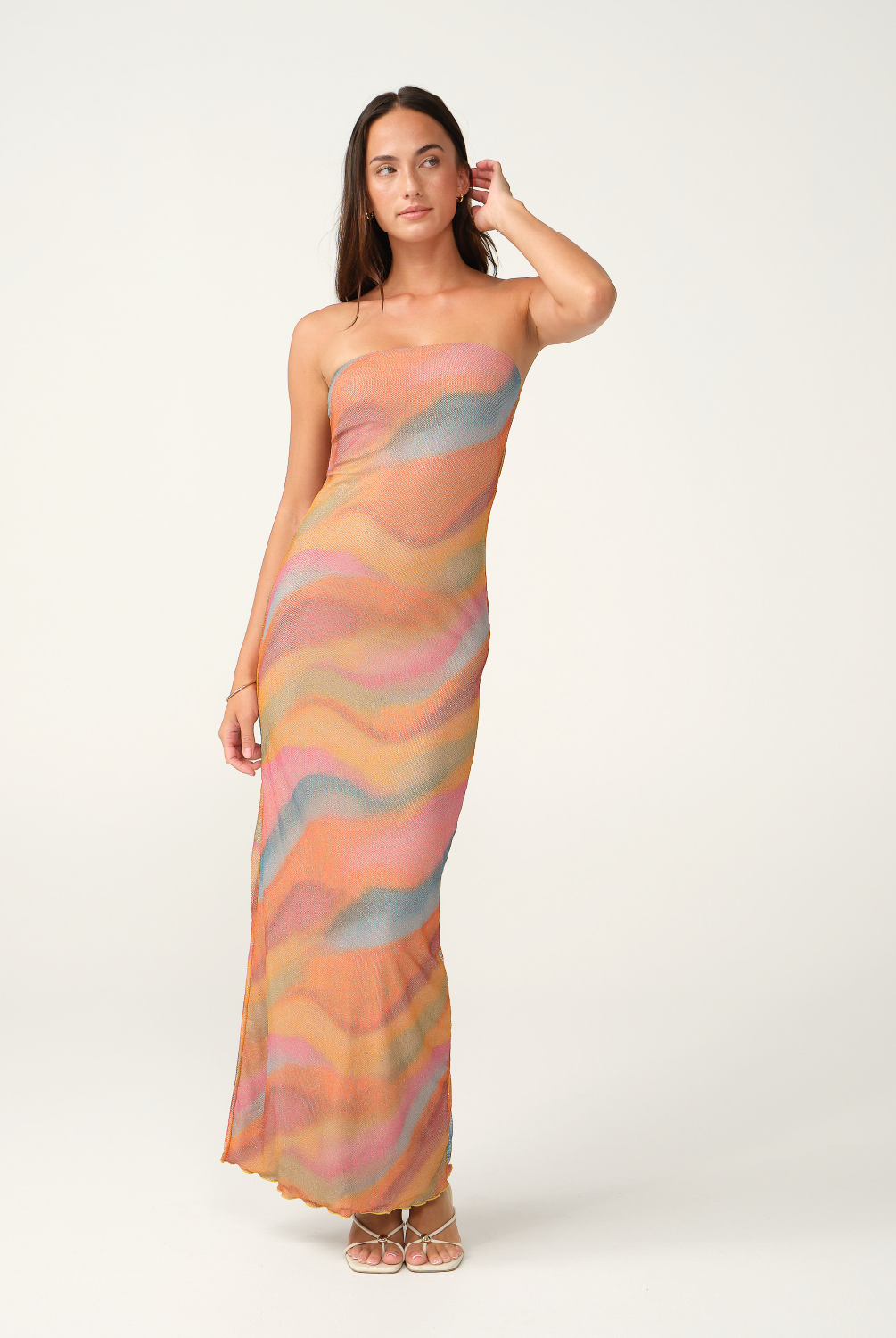 A woman modeling a strapless maxi dress with a wavy, abstract pattern in pastel shades of orange, pink, beige, and light blue, set against a plain light gray studio background.