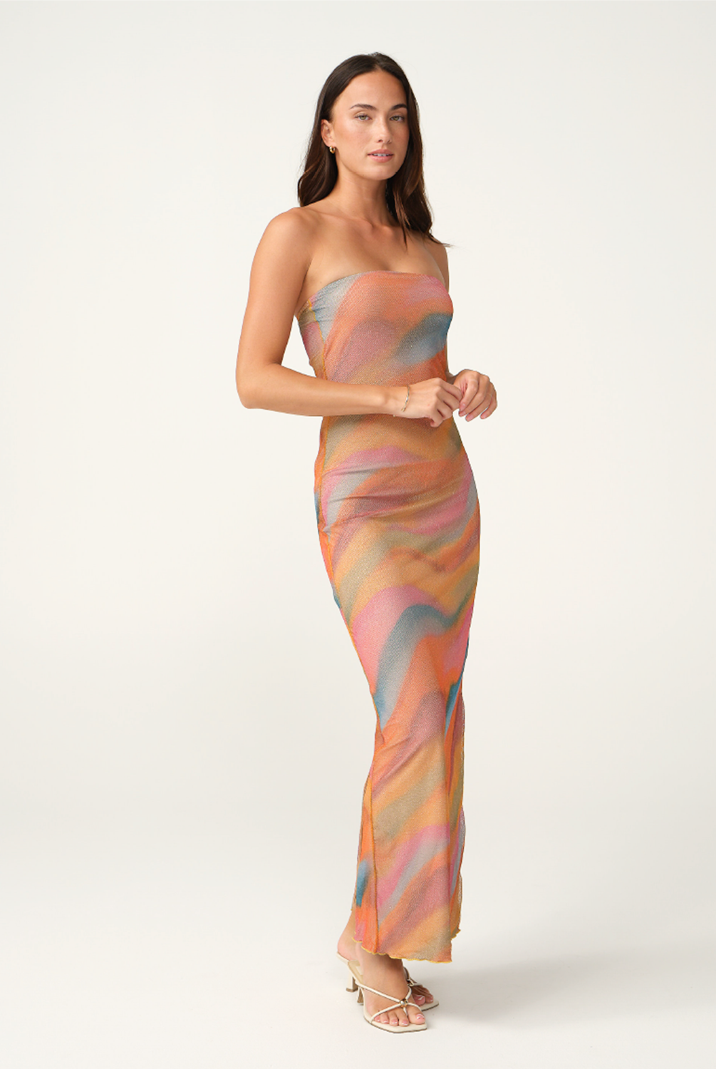 A woman modeling a strapless maxi dress with a wavy, abstract pattern in pastel shades of orange, pink, beige, and light blue, set against a plain light gray studio background.