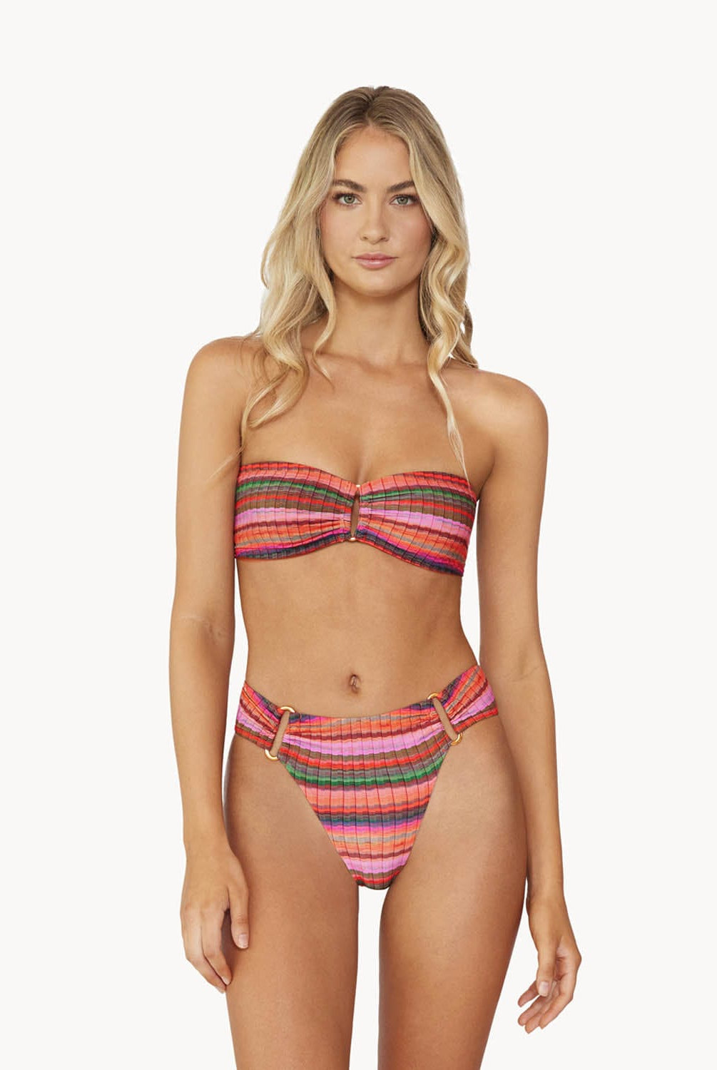 Blonde woman wearing a multi-colored stripe print bandeau bikini with gold details stands in front of white wall.
