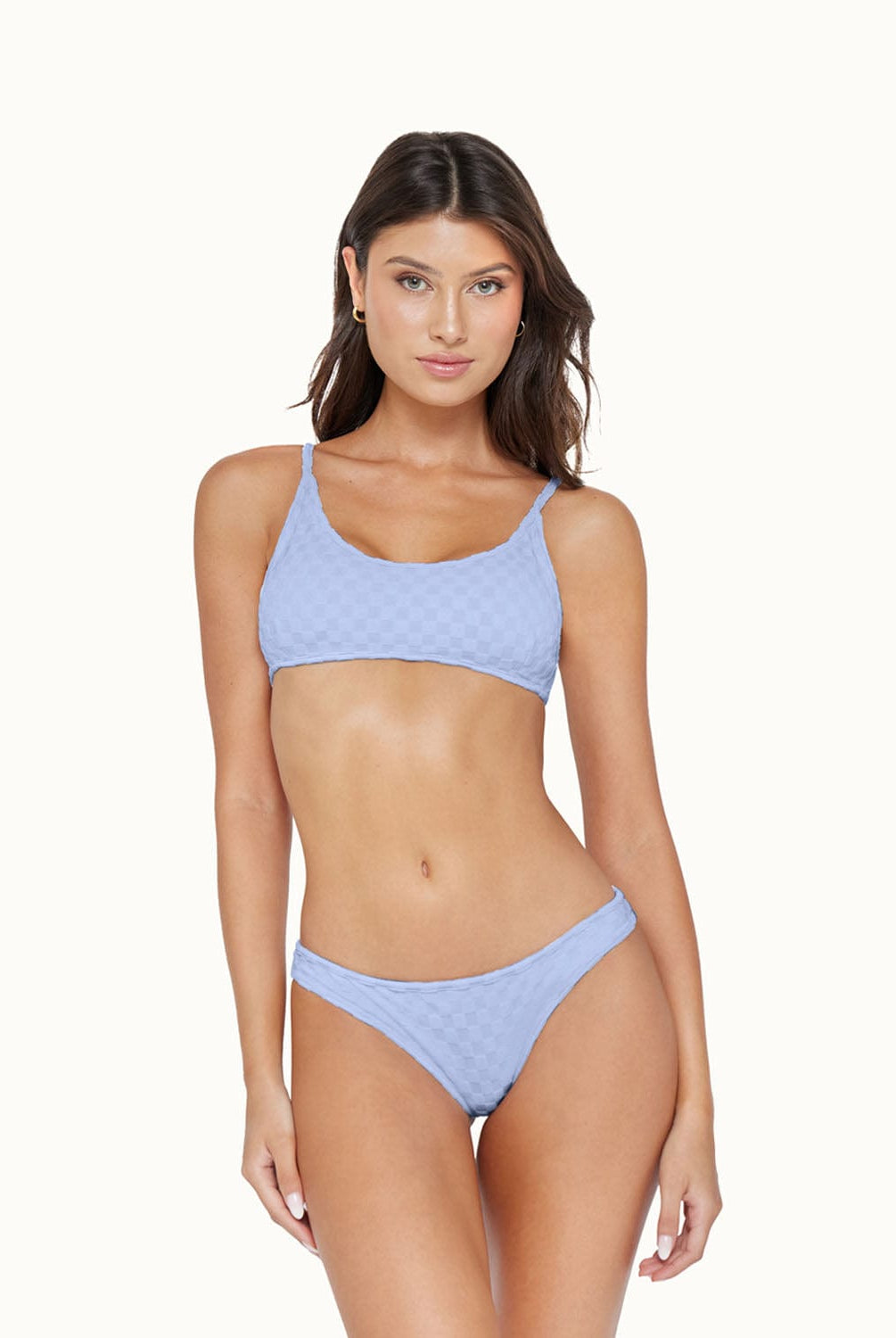 A brunette woman wearing a blue bikini standing in front of a white wall.