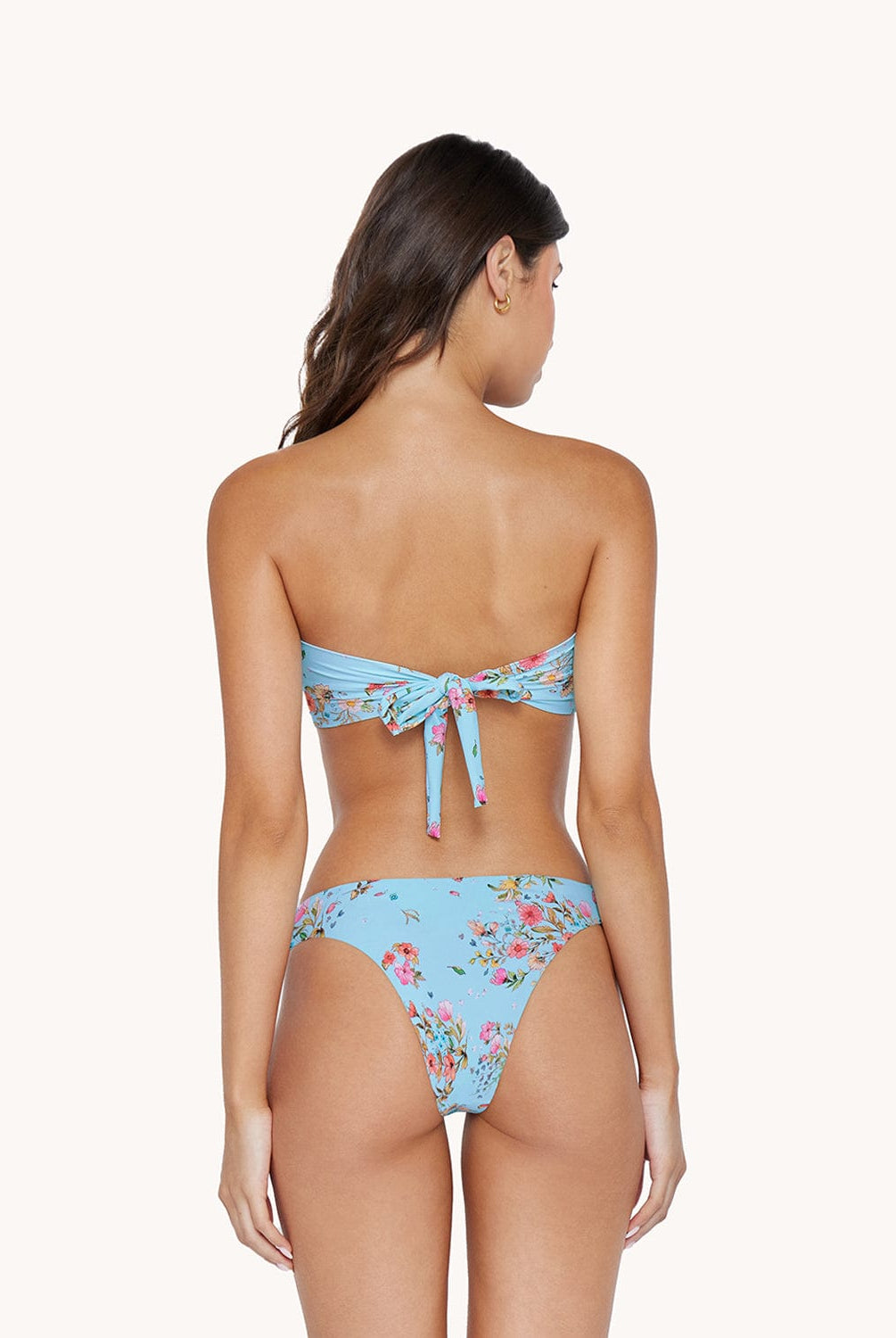 A brunette woman wearing a blue bikini with a floral print facing towards a white walll.