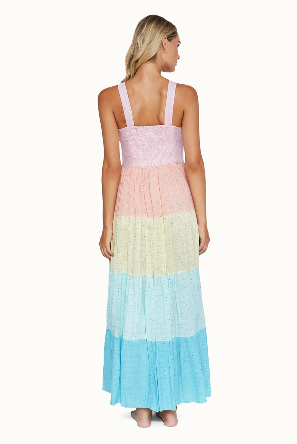 A blonde woman wearing a multi colored dress facing towards a white wall.