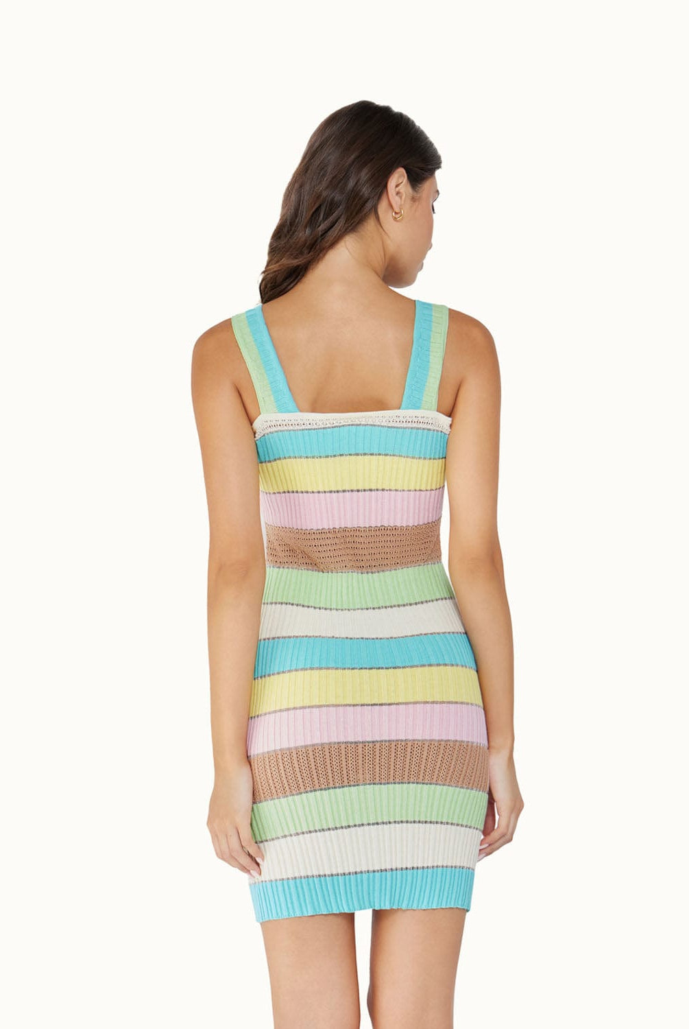 A brunette woman wearing a multi colored dress facing towards a white wall.