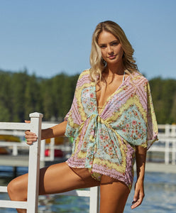 A blonde woman wearing a short floral print cover up standing  on a dock near the water.