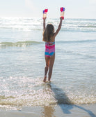 Child in a colorful swimsuit standing in shallow water with arms raised, holding colorful flippers