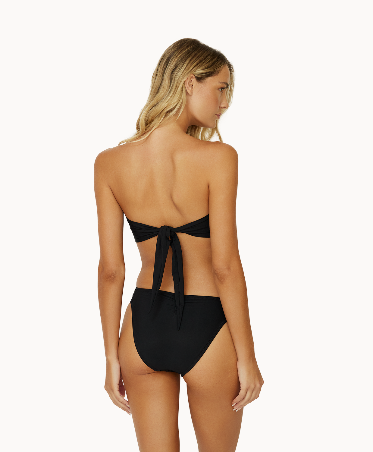 A woman wearing a black bikini facing towards a white wall.