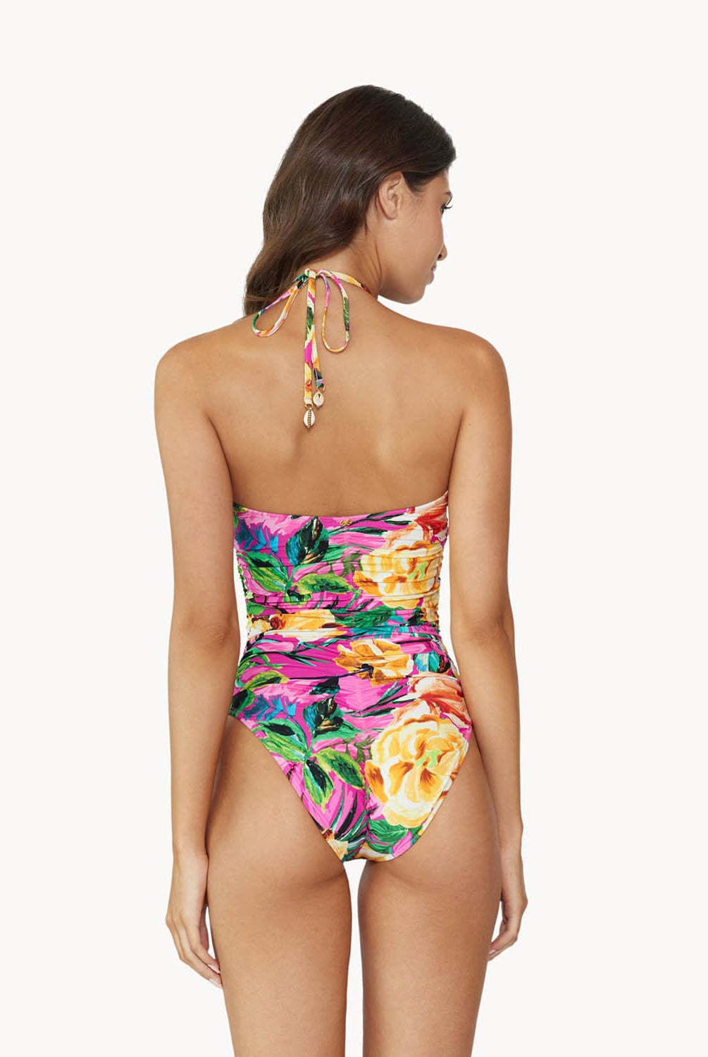 Brunette woman wearing pink tropical print ruched one piece swimsuit facing backwards towards white wall.