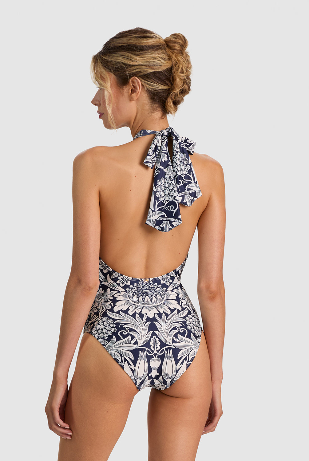 A woman shown from the back wearing a one-piece halter swimsuit with a navy blue and white botanical pattern, set against a solid light gray background.