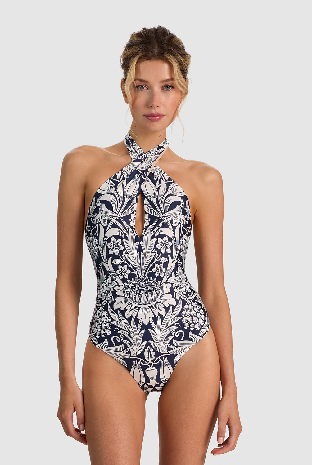 A woman modeling a one-piece halter swimsuit with a navy blue and white botanical pattern, set against a solid light gray background.