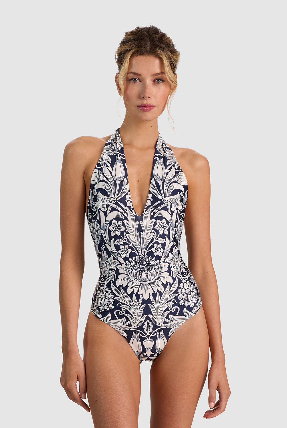 A woman modeling a halter-neck one-piece swimsuit with a navy blue and white baroque floral pattern, set against a solid light gray background.