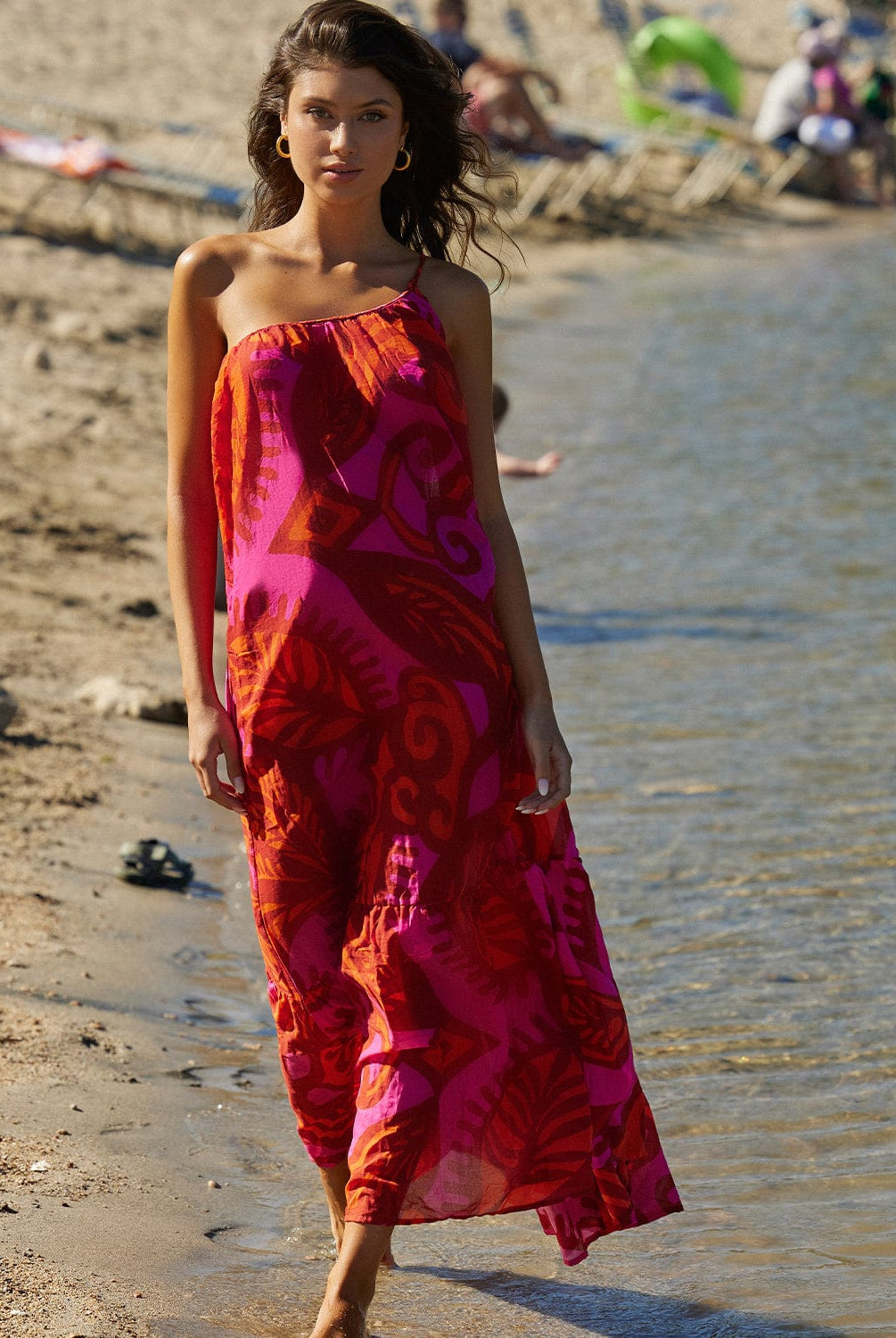 Brunette woman wearing an ankle length dress with one shoulder strap. She is walking on the beach near a lake.