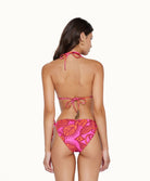 A brunette woman wearing a pink and red bikini facing towards a white wall.