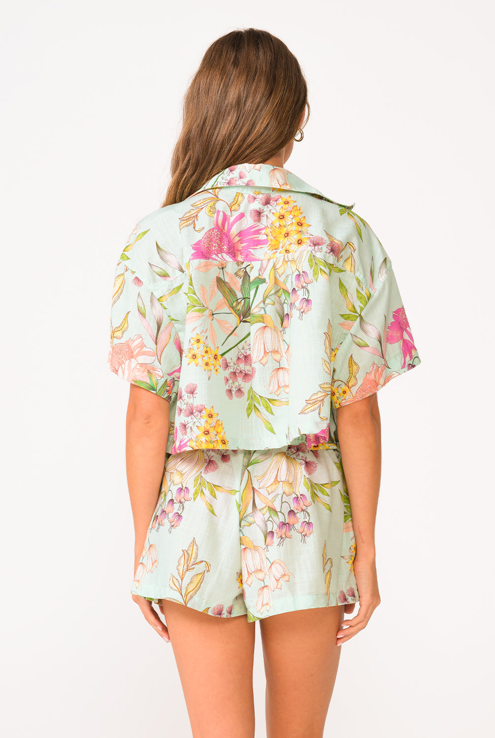 A woman wearing a light blue floral shirt and floral shorts facing towards a white wall.
