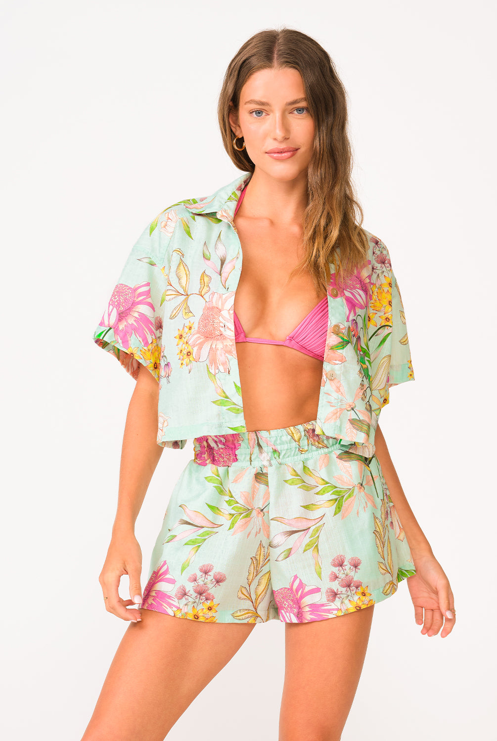 A woman wearing a bright pink halter bikini top and floral shorts standing against a white wall.