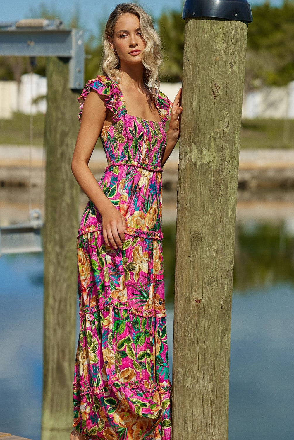 Blonde woman wearing a maxi pink tropical dress with sequins stands on a dock by water.