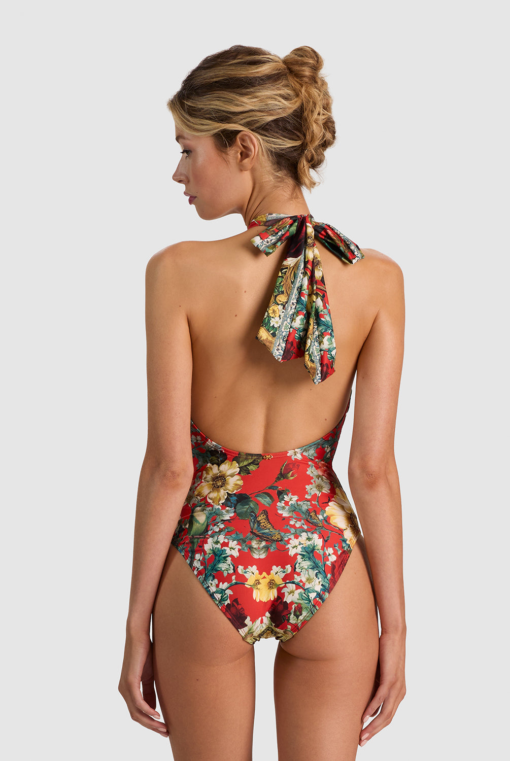 A woman shown from the back wearing a one-piece swimsuit with a red base and a colorful floral print, featuring a tie-neck design. The background is a solid light gray studio backdrop.