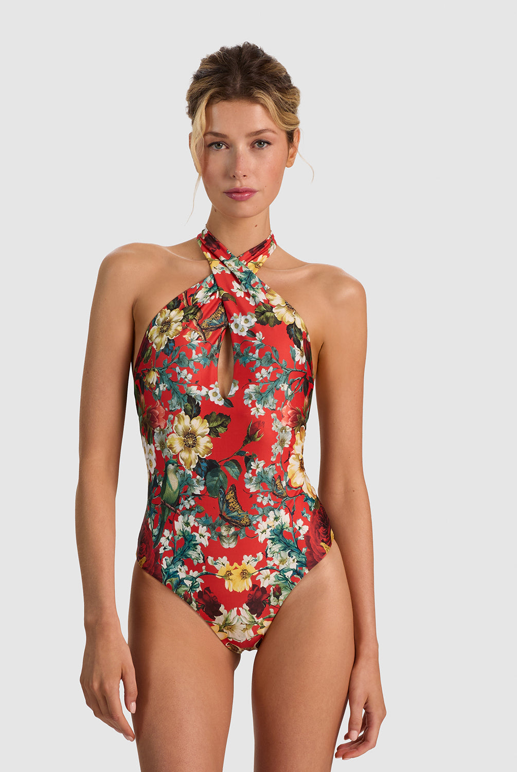 A woman modeling a red one-piece swimsuit with a vibrant floral print in yellow, white, and green, set against a solid light gray background.