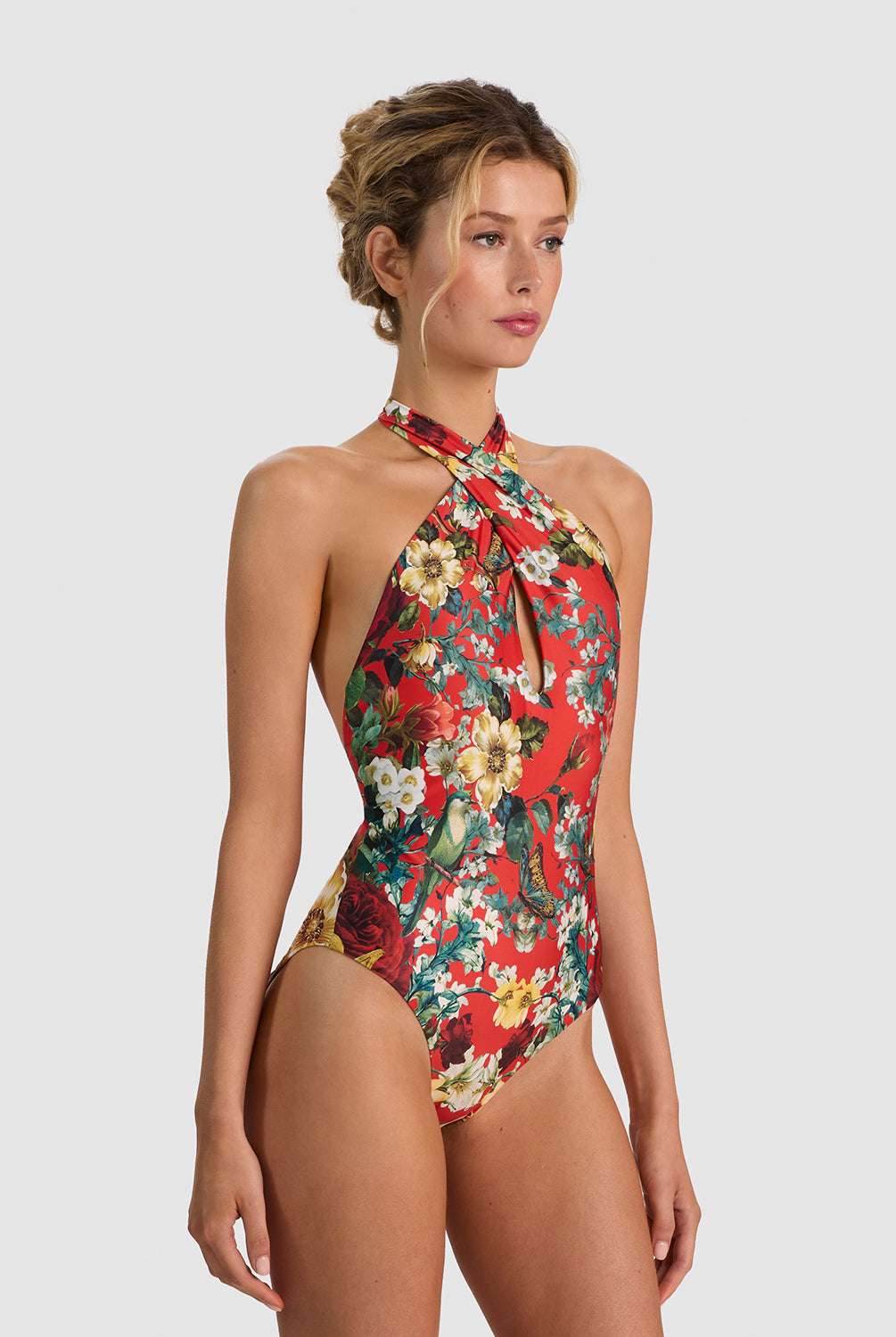 A woman modeling a one-piece halter swimsuit with a vibrant red, green, and yellow floral pattern against a solid light gray background.