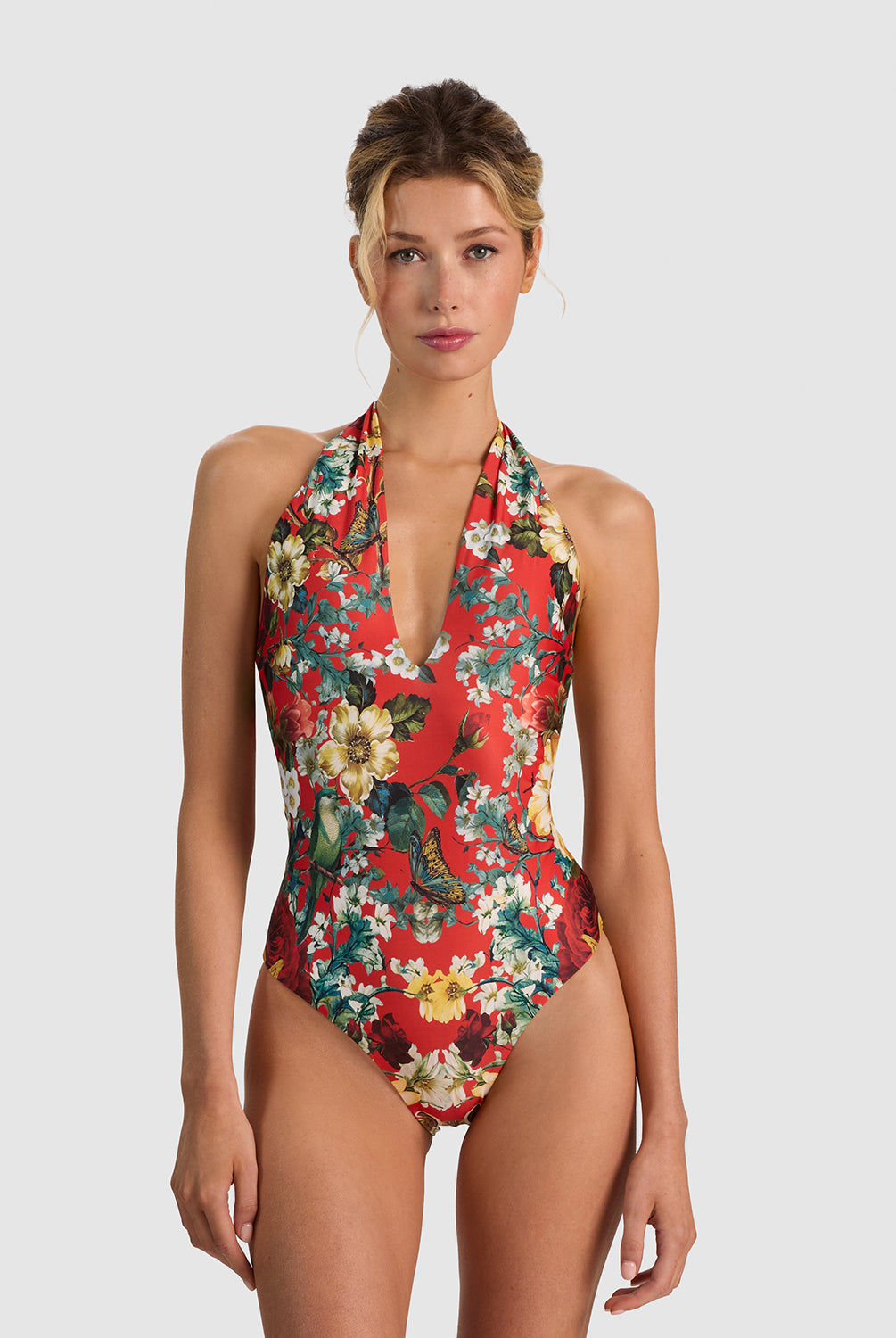 A woman modeling a red one-piece halter swimsuit with a colorful floral and bird print, set against a solid light gray background.