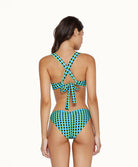 A brunette woman wearing a blue and green checkered bikini facing towards a white wall.