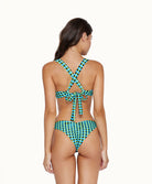 A brunette woman wearing a blue and green checkered bikini facing towards a white wall. 