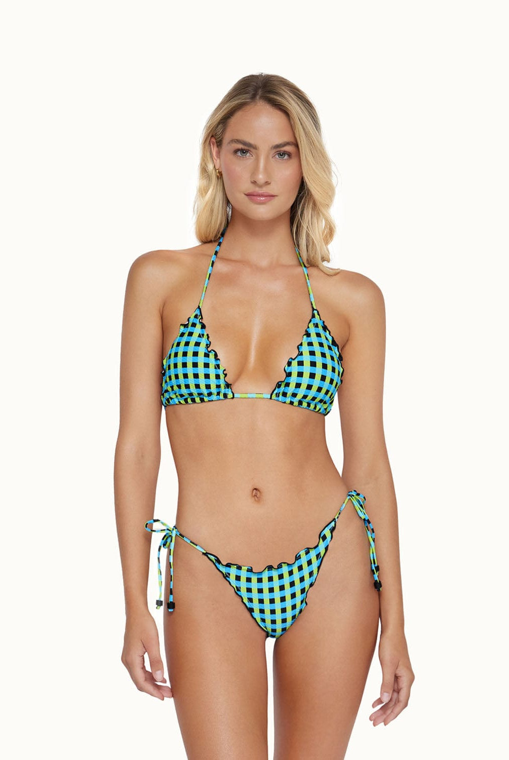A blonde woman wearing a blue and green checkered bikini stands against a white wall. 