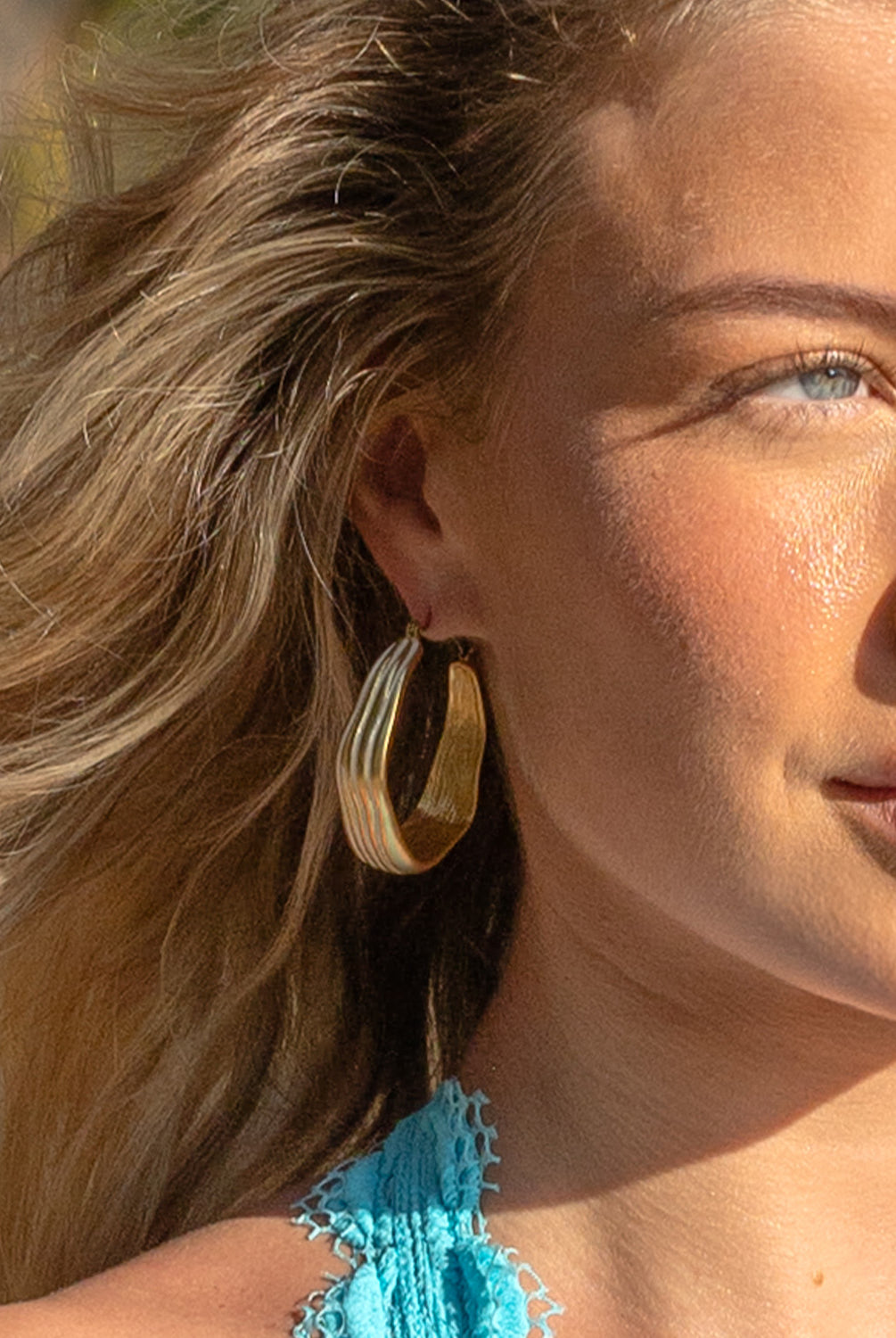 A woman wearing a teal bikini top and gold hoop earrings standing outside near palm trees.