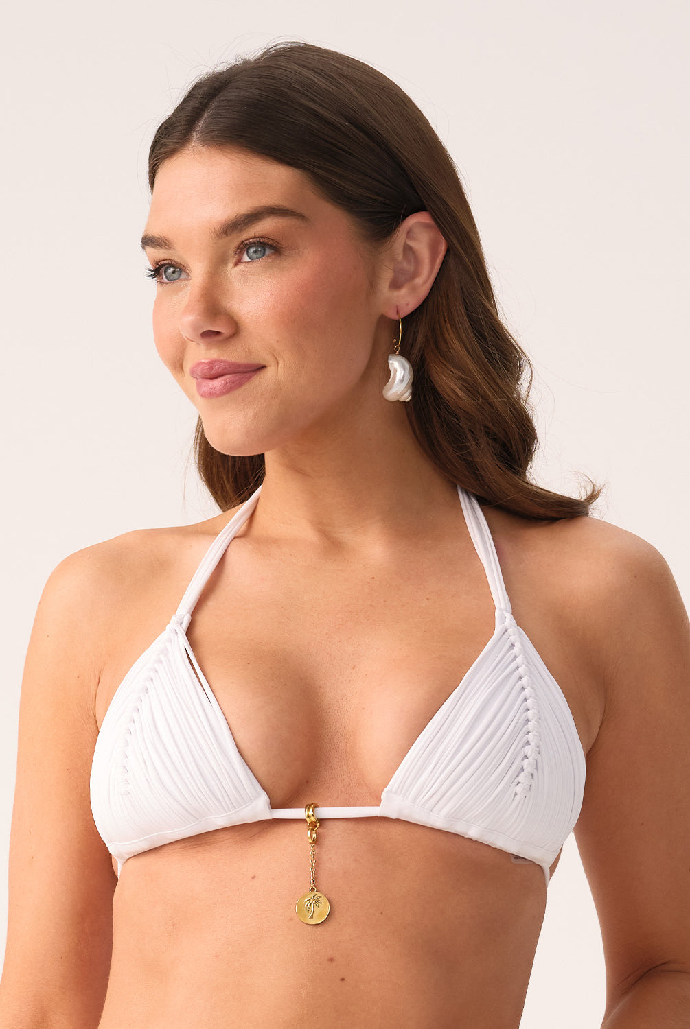 A woman wearing a white triangle top bikini and shell earrings standing against a white wall.