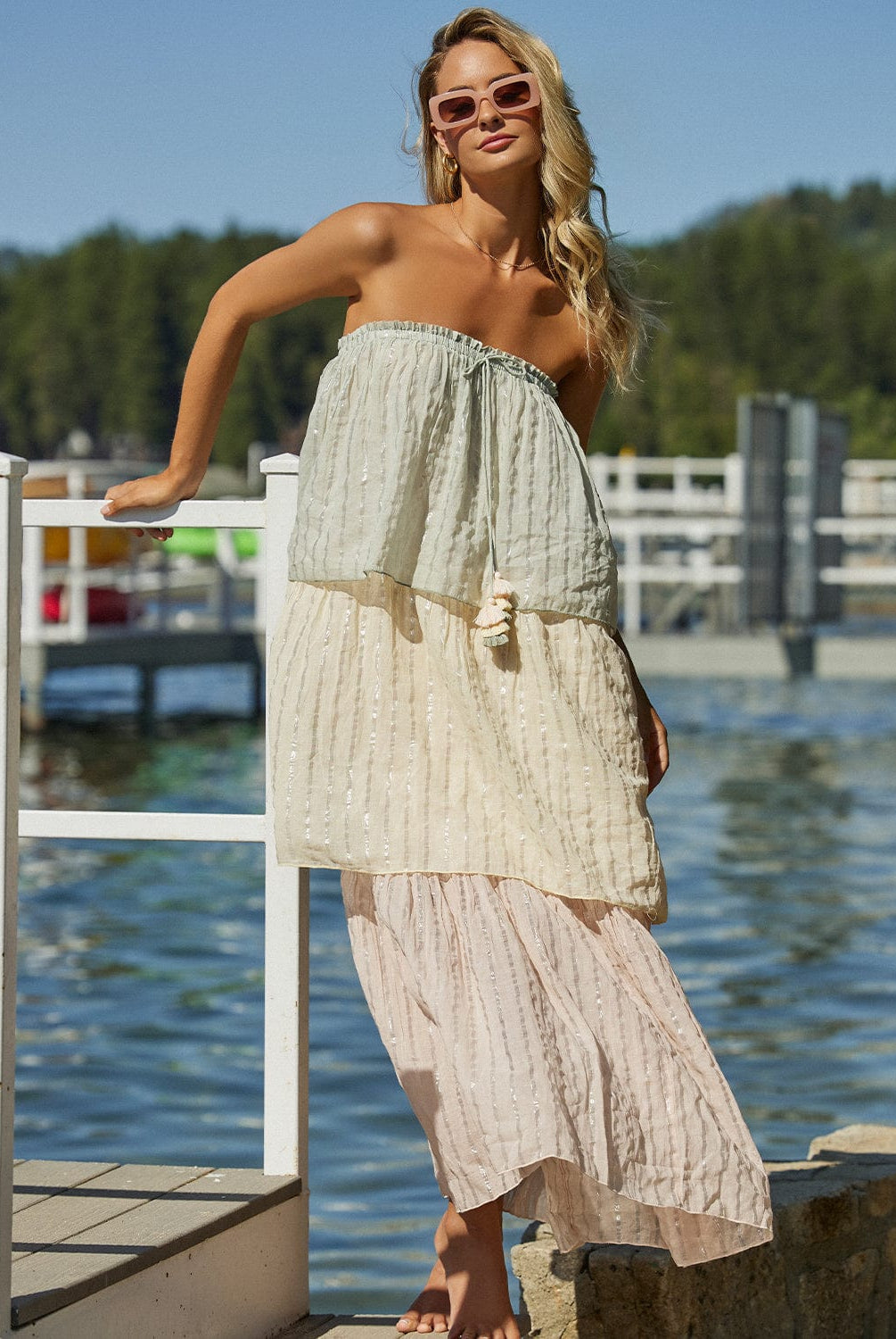 A blonde woman wearing a multi colored floor length dress leaning against a dock near the water.