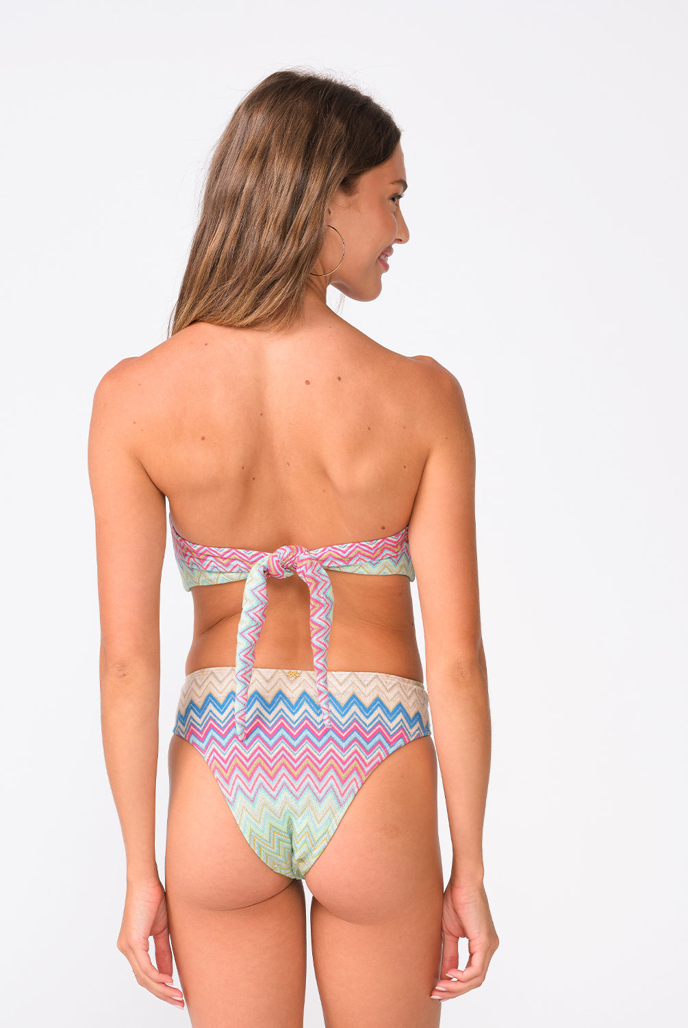 A woman wearing a colorful striped bandeau bikini facing towards a white wall.