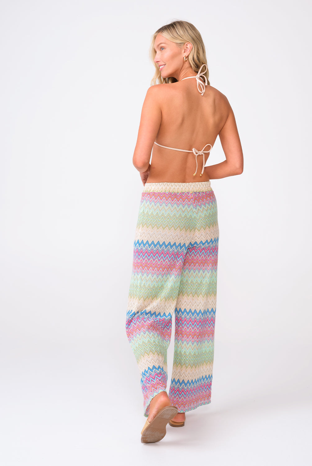 A woman wearing a striped triangle top and matching pants facing towards a white wall.