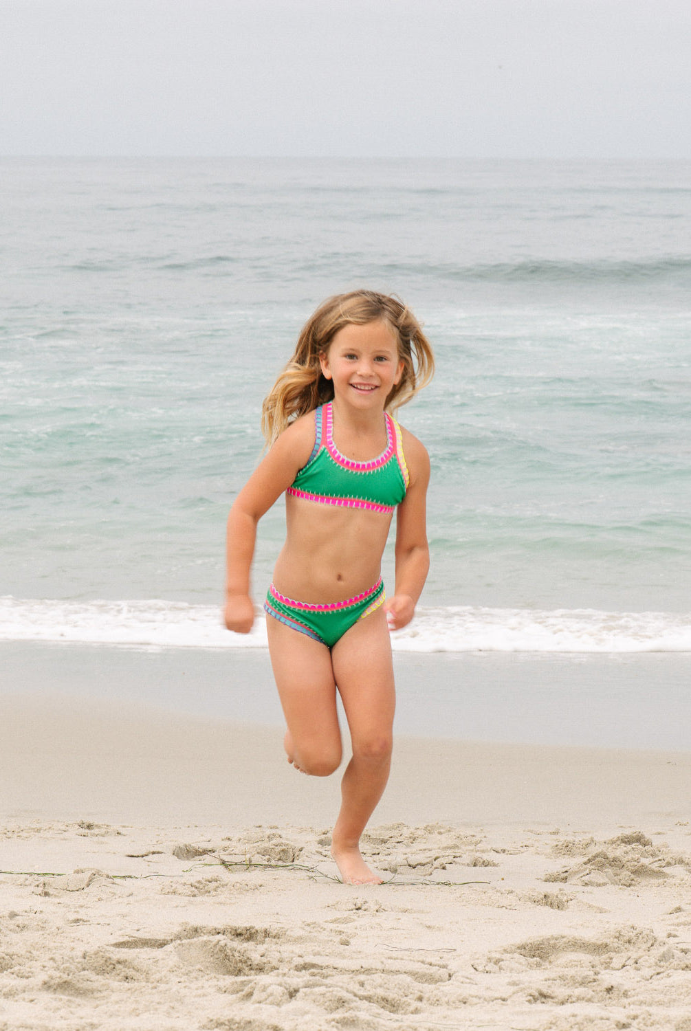 Young girl in a green bikini with pink trim standing on a beach.