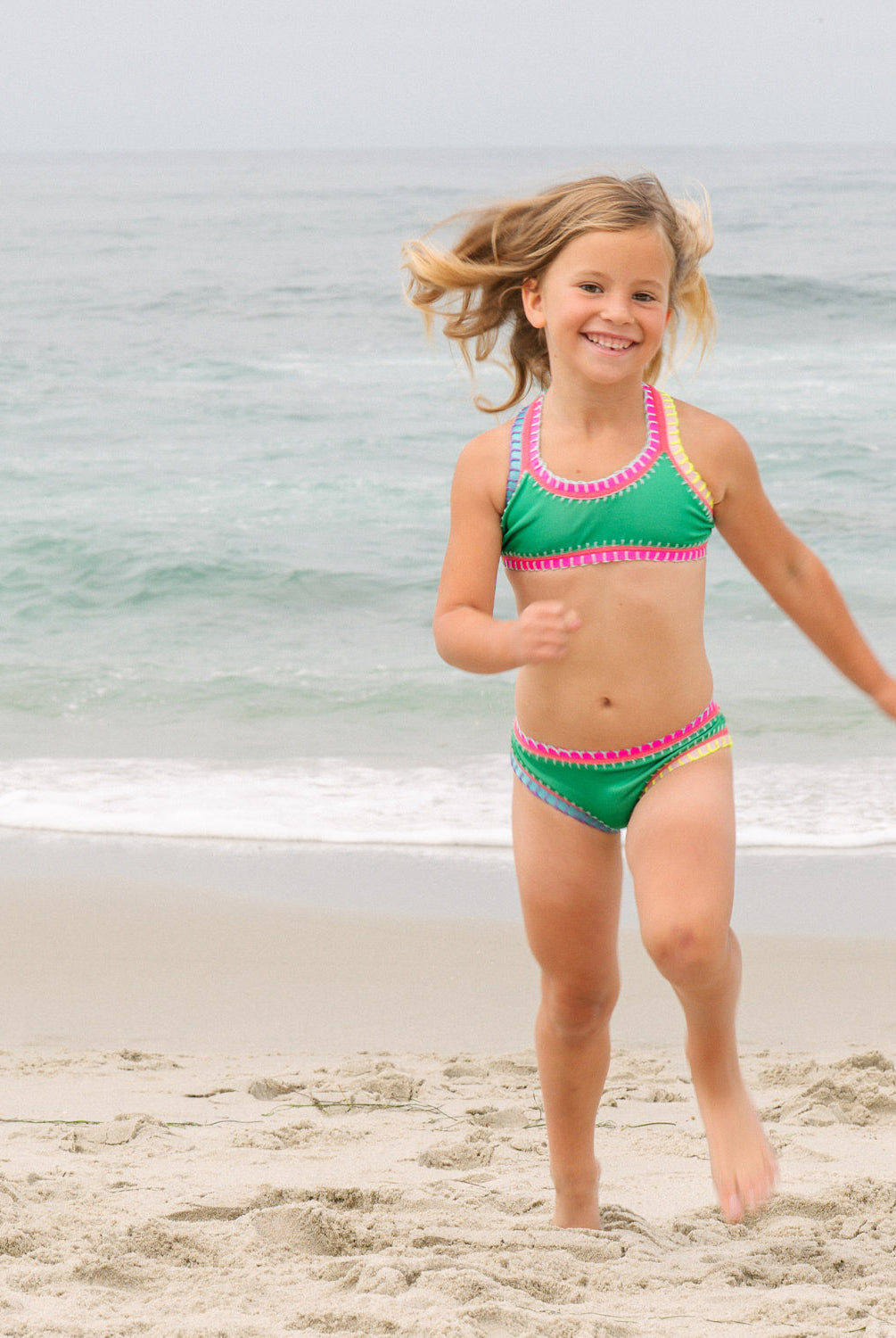 Young girl in a green and pink swimsuit running on a beach with ocean in the background