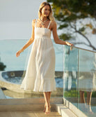 A blonde woman wearing a midi length white dress standing outside near a glass wall.