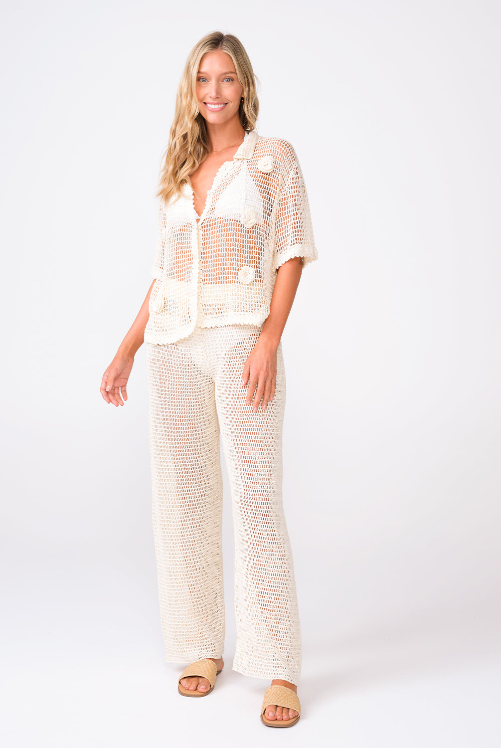 A woman wearing an ivory knit shirt and matching pants standing against a white wall.