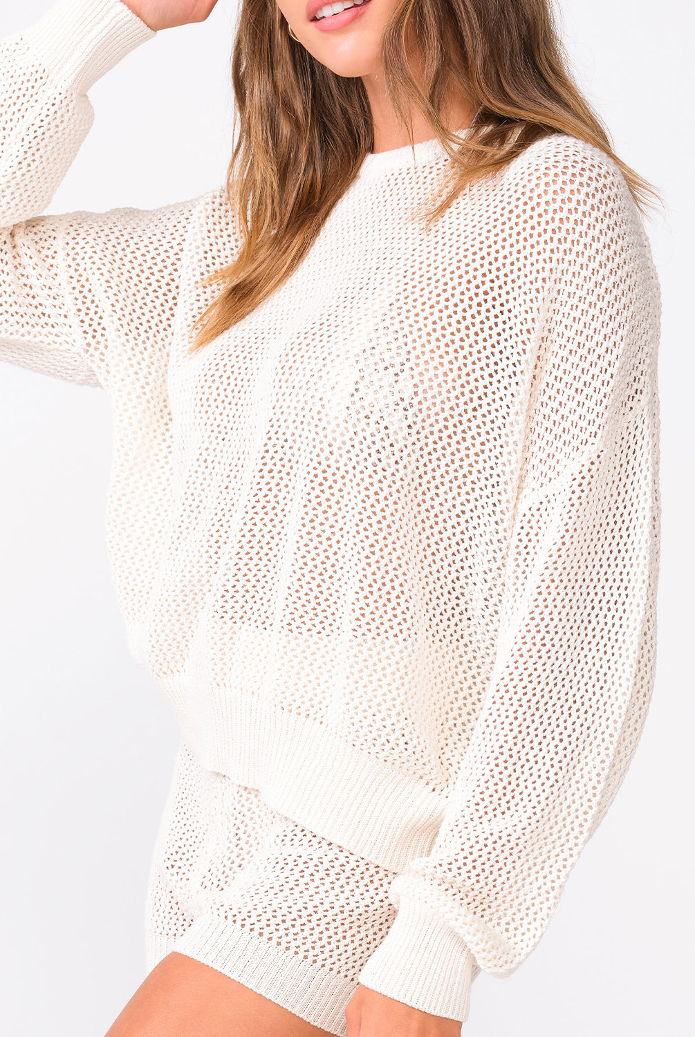 A woman wearing an ivory knit sweater and matching shorts standing against a white wall.