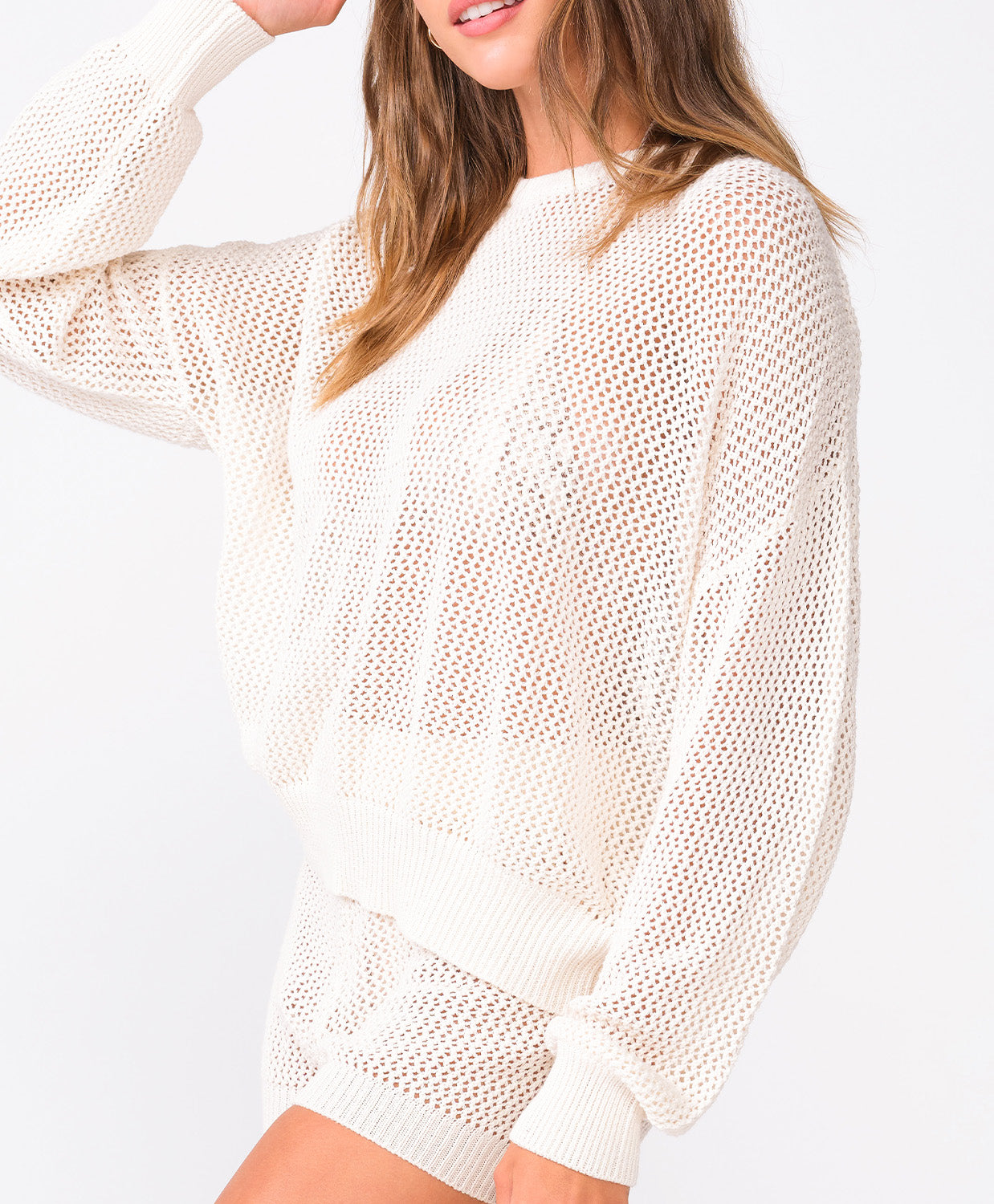 A woman wearing an ivory knit sweater and matching shorts standing against a white wall.