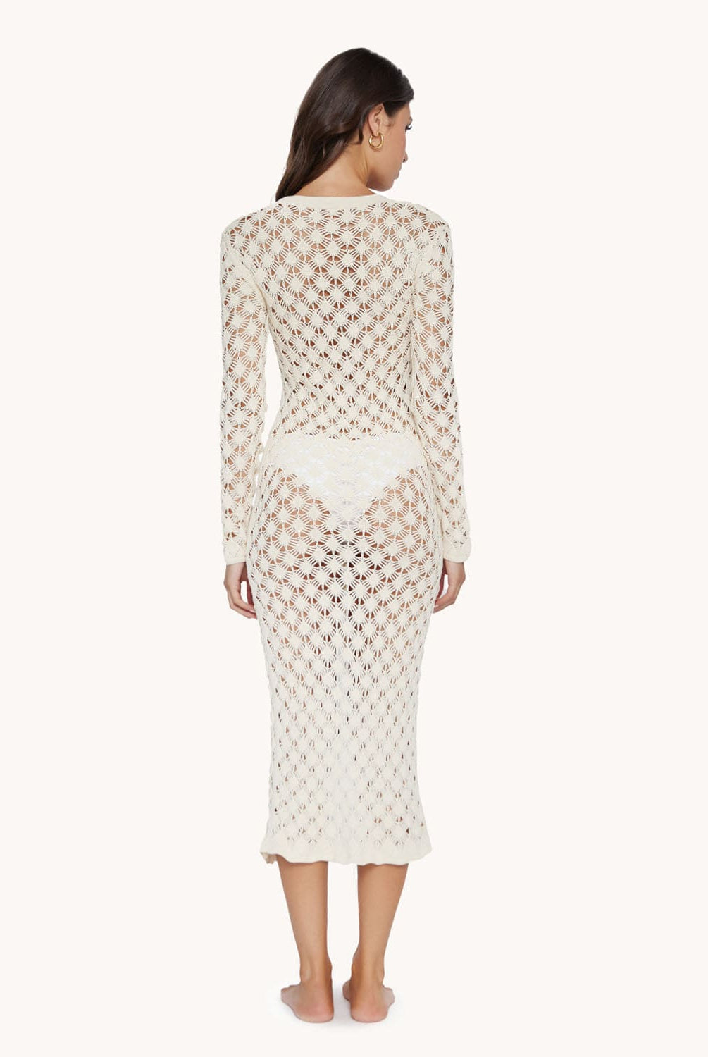 A brunette woman wearing a white bikini and Ivory long sleeve mesh cover up facing towards a white wall.