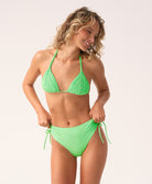 A woman wearing a green triangle top bikini and matching high waist bottom standing against a white wall.