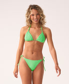 A woman wearing a green triangle top bikini and matching tie-side bottom standing against a white wall.