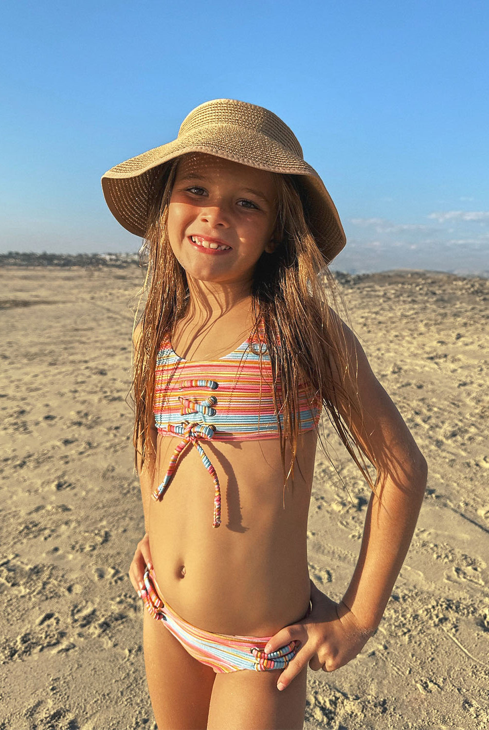 A young girl wearing a striped bikini and bucket hat standing on a beach.