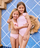 One girl is wearing a light pink bikini and another girl is wearing a light pink one piece with a bow.