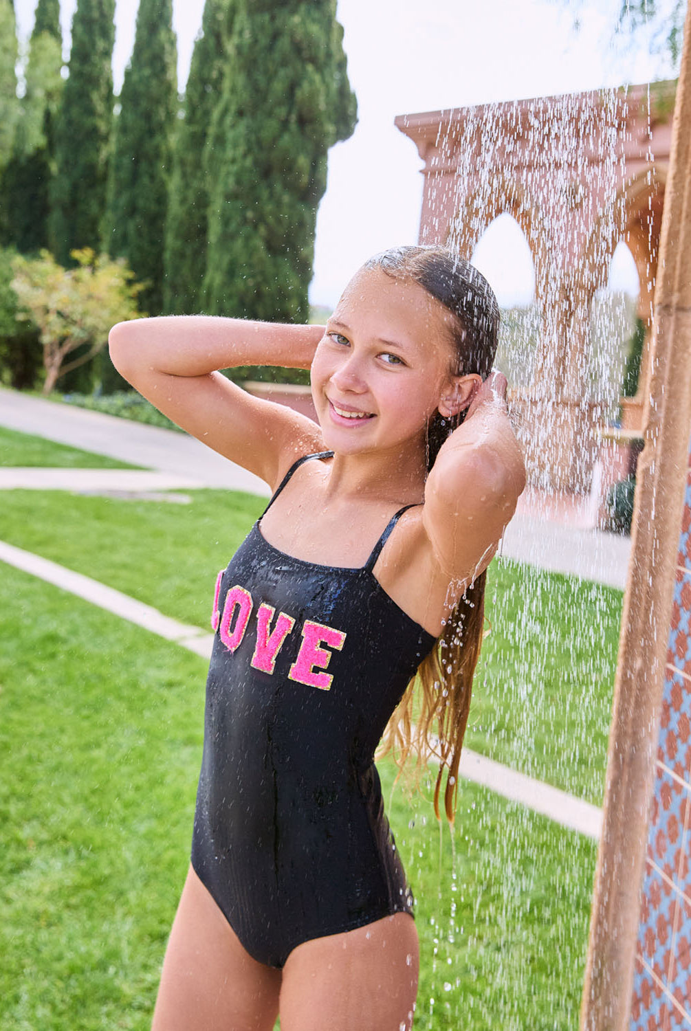 A brunette girl wearing a black one piece swimsuit standing under a shower.