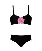 A black bikini with a pink rose on the top against  white wall.