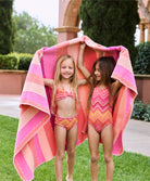 A blonde girl wearing a pink and orange striped bikini and a brunette girl wearing an orange and pink striped one piece. Both girls are standing on the grass outside.