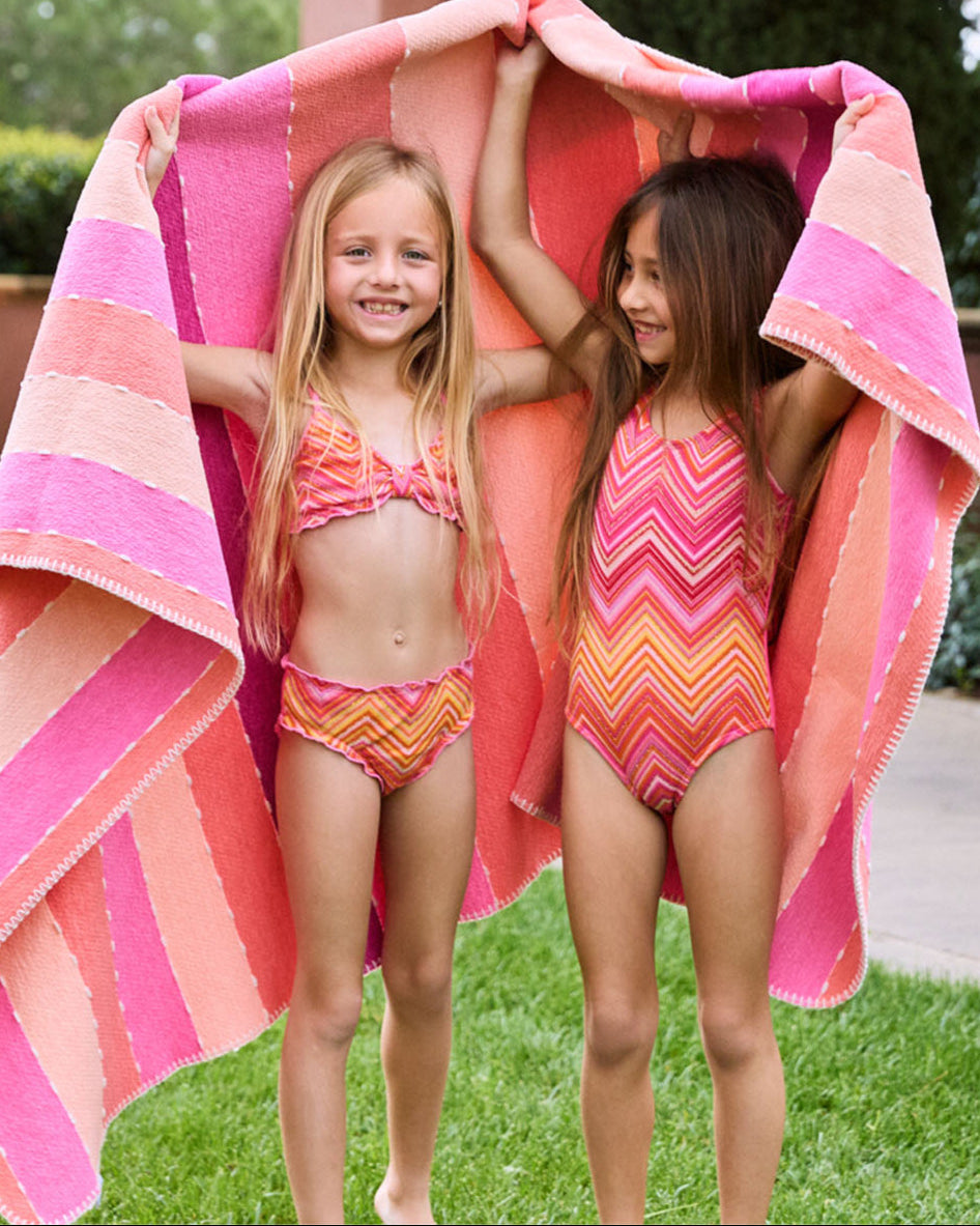 A blonde girl wearing a pink and orange striped bikini and a brunette girl wearing an orange and pink striped one piece. Both girls are standing on the grass outside.